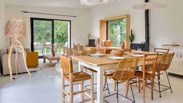 A modern dining area with a wooden table, wicker chairs, and a large window offering a view of greenery. - Casa Sitha magnifique maison de vacances de 110m2 (Gouville-sur-Mer)