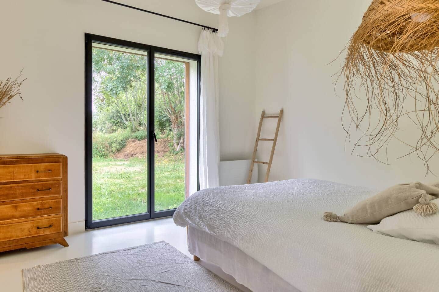 A bedroom with a bed, a wooden dresser, a large window with a view of greenery, and a ladder against the wall.
