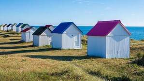 A row of beach huts with corrugated metal roofs and white walls, aligned along a grassy beach with the sea in the background. - Casa Sitha magnifique maison de vacances de 110m2 (Gouville-sur-Mer)