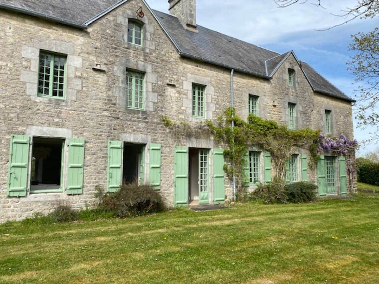 Un bâtiment en pierre avec des volets verts, une pelouse verdoyante et un ciel dégagé.