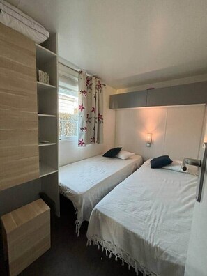 A small room with two beds, a shelf unit, and a window with curtains. - Mobilhome in Residential leisure park (Hauteville-sur-Mer)