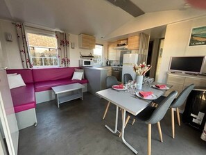 A compact living space with a sofa, dining table, chairs, kitchenette, and a television. - Mobilhome in Residential leisure park (Hauteville-sur-Mer)