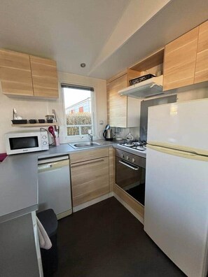 A compact kitchen with a microwave, stove, sink, and wooden cabinets. - Mobilhome in Residential leisure park (Hauteville-sur-Mer)