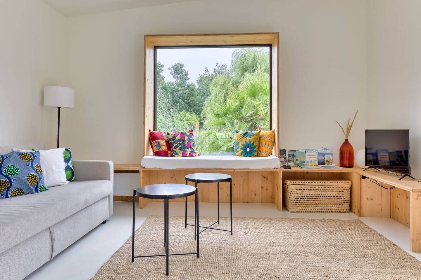 Una sala de estar moderna con un sofá, dos mesas redondas, un ventanal con vistas a zonas verdes, un televisor y una estantería.