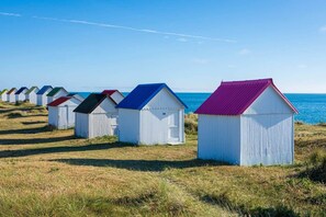 Eine Reihe von Strandhütten mit wellenförmigen Metall-Dächern und weißen Wänden, die an einem grasbewachsenen Strand entlang verlaufen, mit dem Meer im Hintergrund.