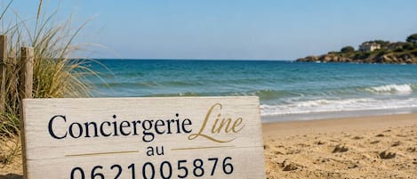 Un panneau en bois indiquant « Conciergerie, appelez le 06 21 00 58 76 », placé sur une plage de sable parsemée de coquillages.
