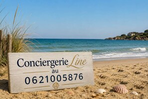 A wooden sign with 'Conciererie Line au 0621005876' on a beach with sand and shells. - Casa ZaO Jolie maison en bois brut de 50m2 à (Gouville-sur-Mer)