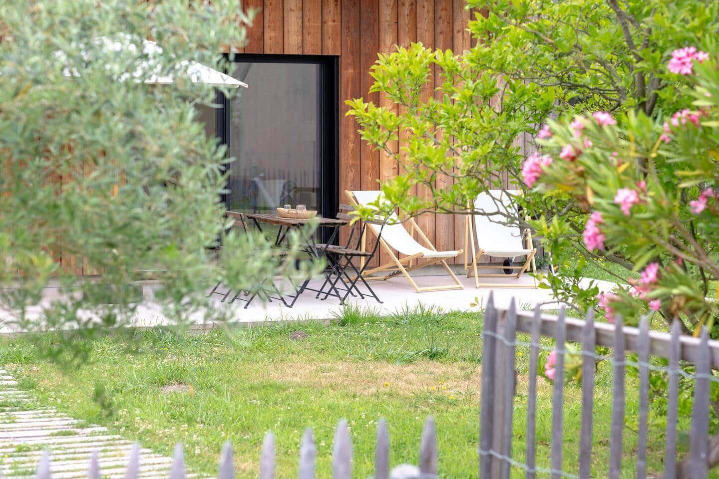 A wooden building with a glass door, outdoor seating with chairs and a table, and a fenced garden area.