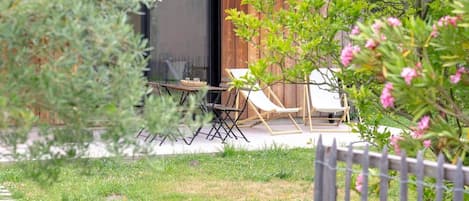 Un edificio de madera con puerta de vidrio, mobiliario exterior con sillas y mesa, y un jardín vallado.