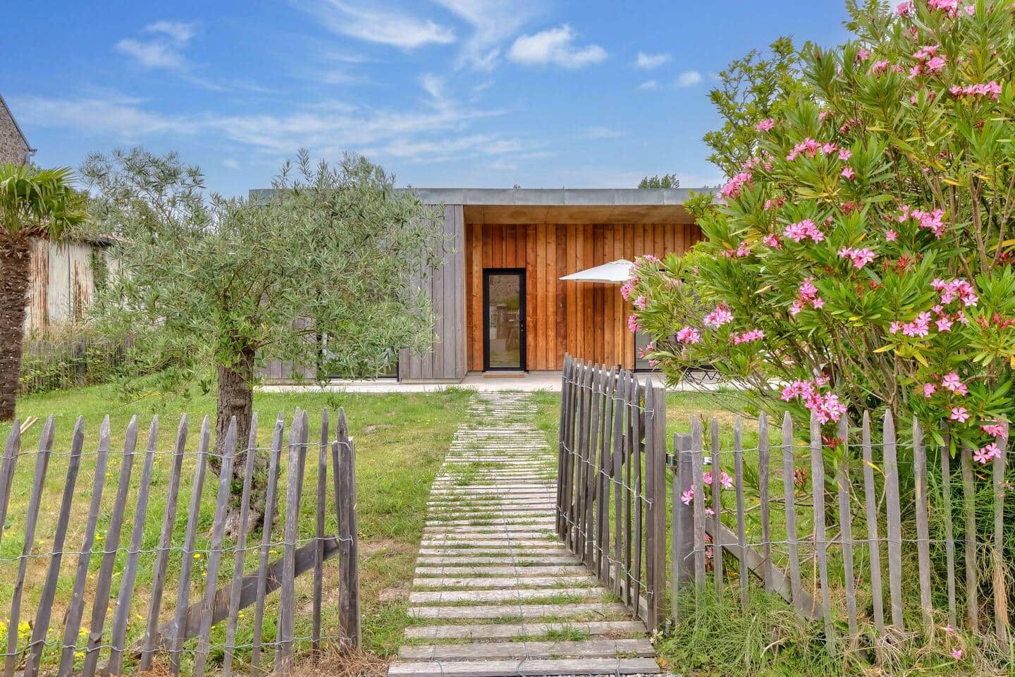 A pathway leading to a modern building with a wooden door, surrounded by a wooden fence and greenery.