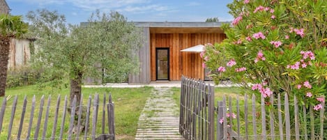 A pathway leading to a modern building with a wooden door, surrounded by a wooden fence and greenery.