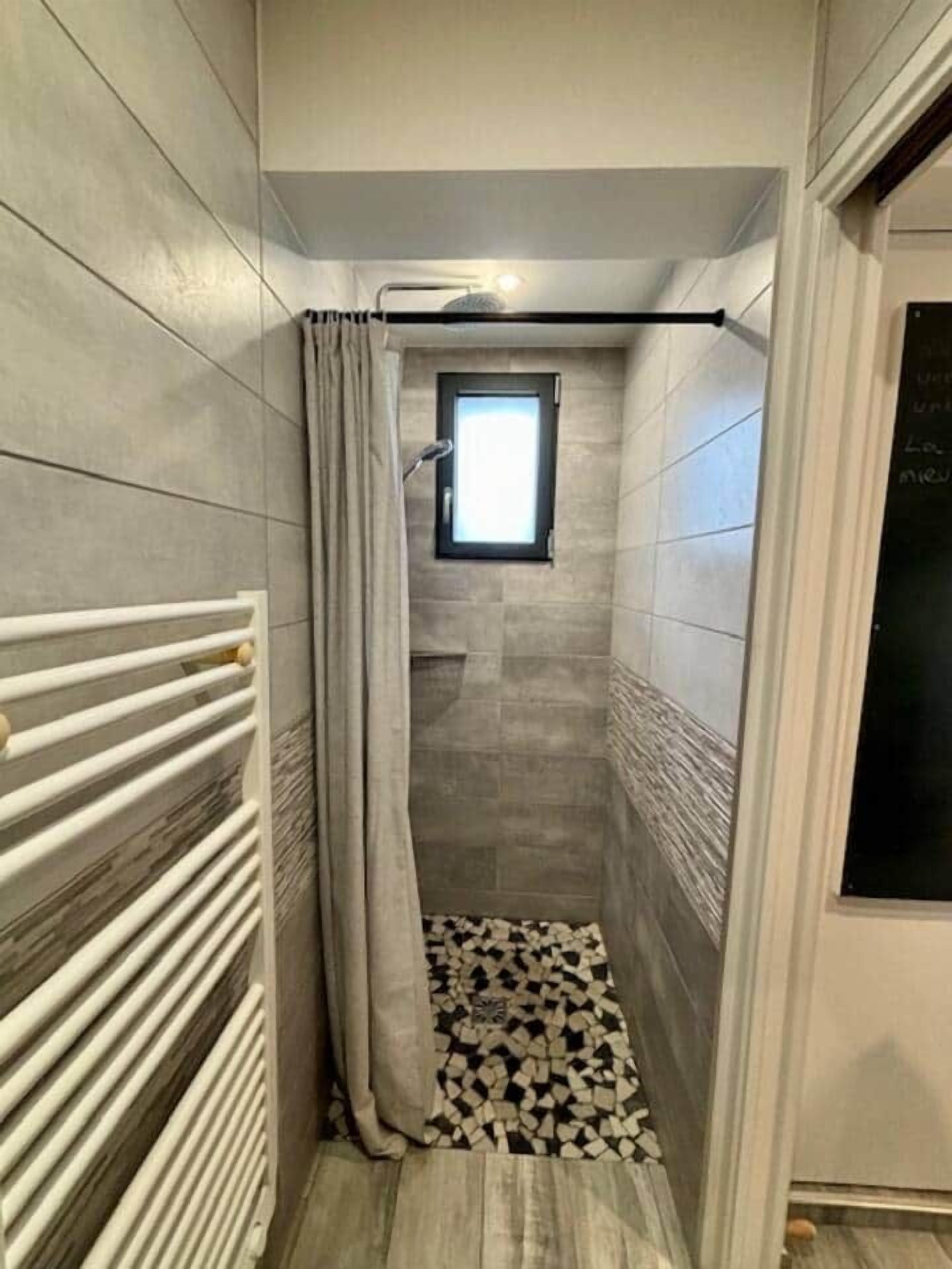 A narrow bathroom with a shower, a window, and a radiator.