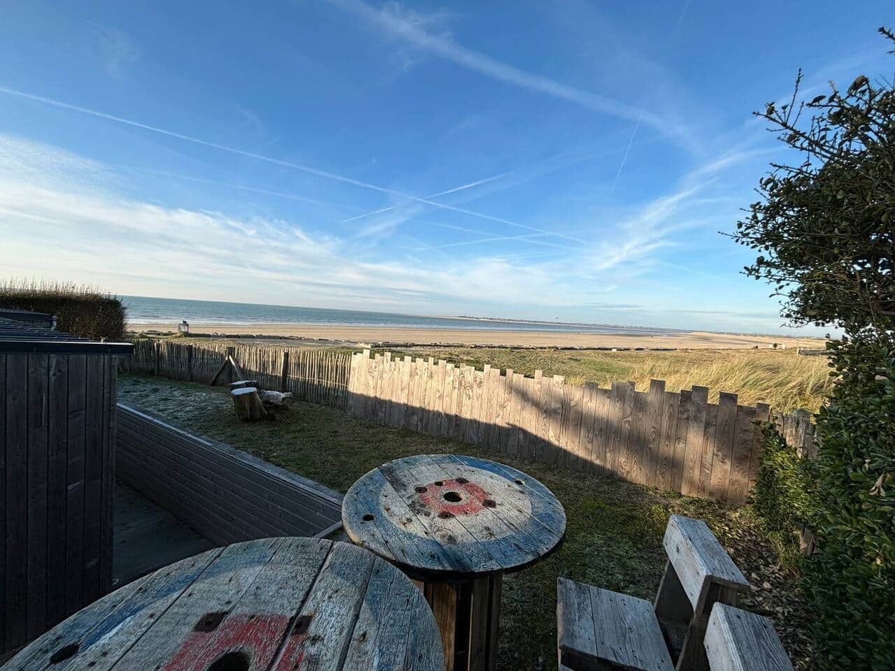 Una terraza de madera con una mesa y sillas, con vista a una playa y campos cubiertos de hierba.