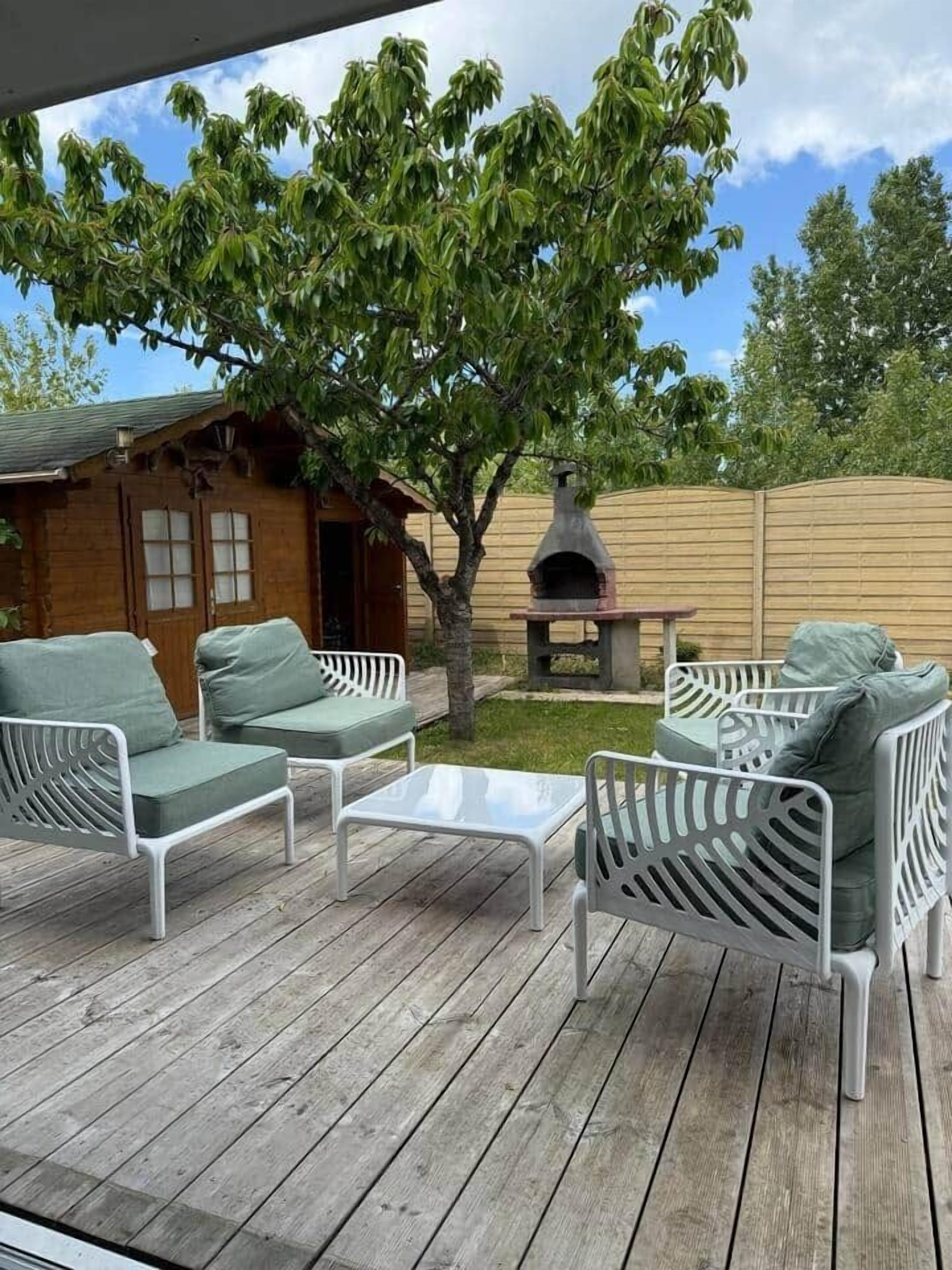 Una terraza de madera con mobiliario exterior, un árbol y una zona de barbacoa.