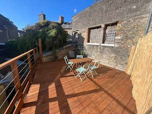 Eine Holzterrasse mit Gartenmöbeln, ein Steinhaus und ein klarer, blauer Himmel.