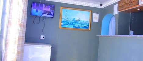 A room with a television mounted on the wall, a framed picture of a cityscape, a white refrigerator, and a counter with a sink.