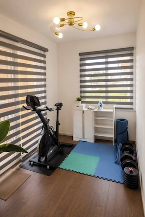A compact room with a stationary bike, a small desk, a shelf, and a window with blinds. - Nicholas Residence (Silang)