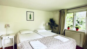 Ein Schlafzimmer mit Doppelbett, Nachttisch, Lampe, gerahmtem Bild an der Wand, Fenster mit Vorhängen und einer Topfpflanze.