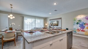 A modern kitchen with a granite countertop, stainless steel sink, and a dishwasher. There is a dining area with a table and chairs, and a large window with curtains. - 5BR Formosa Villa Near Disney | Private Pool (Kissimmee)