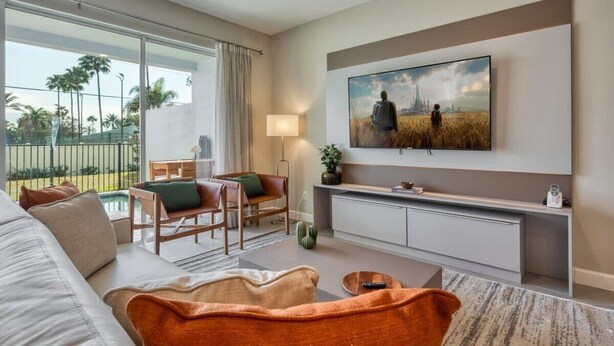 A modern living room with a large TV, a sofa, a dining area, and a view of palm trees through the window. - 5BR Formosa Villa Near Disney | Private Pool (Kissimmee)