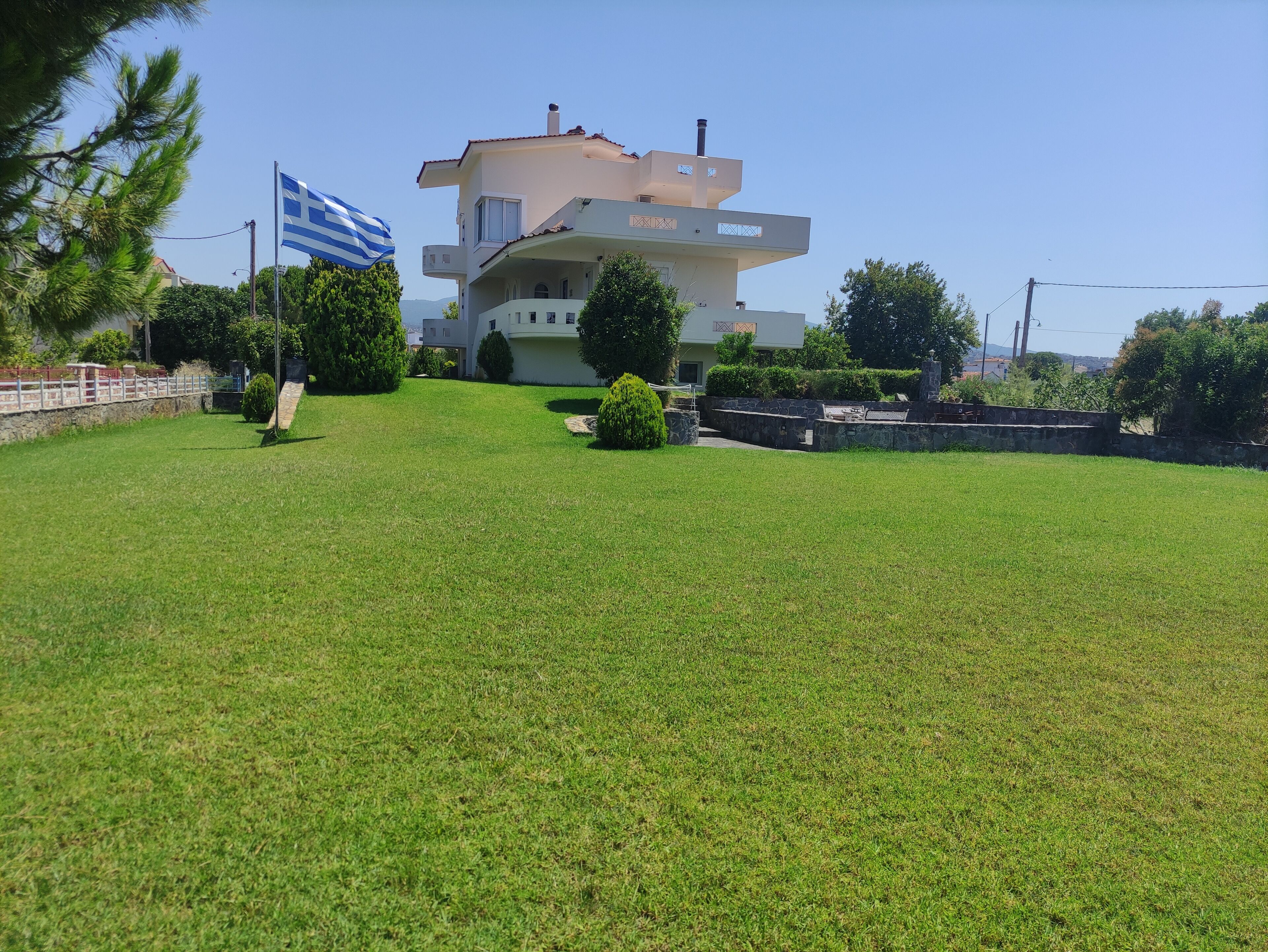 Una moderna casa a due piani con tetto piatto e un balcone, immersa in un prato curato e circondata da un muro in pietra.