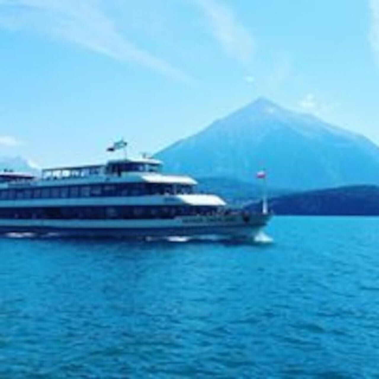 Un navire de croisière doté de plusieurs ponts, arborant un pavillon et ayant une chaîne de montagnes en toile de fond.