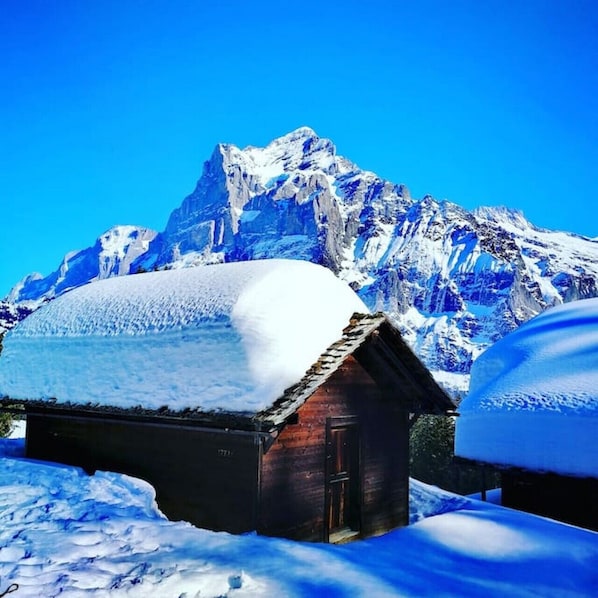Une cabane en bois dont le toit est recouvert de neige, entourée d’un paysage enneigé et avec une montagne en arrière-plan.