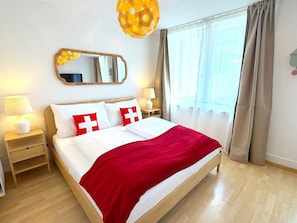 Une chambre d’hôtel avec un lit, une table de chevet, une lampe, un miroir et une fenêtre avec des rideaux.