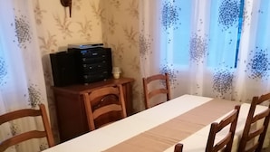 Zona de menjador amb una taula llarga, cadires de fusta, un petit armari i cortines amb dibuixos.