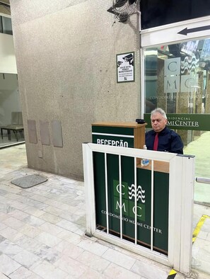 Un mostrador de recepción con un letrero del Condomínio MMI Center, un letrero de cámara de seguridad y una puerta de entrada de vidrio.