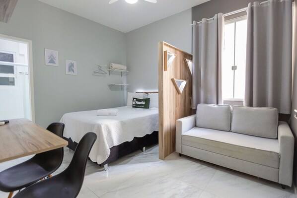 Ein Zimmer mit einem Bett, einem Schlafsofa, einem Tisch und Stühlen. Zwei an der Wand befestigte Regale mit Gegenständen darauf.