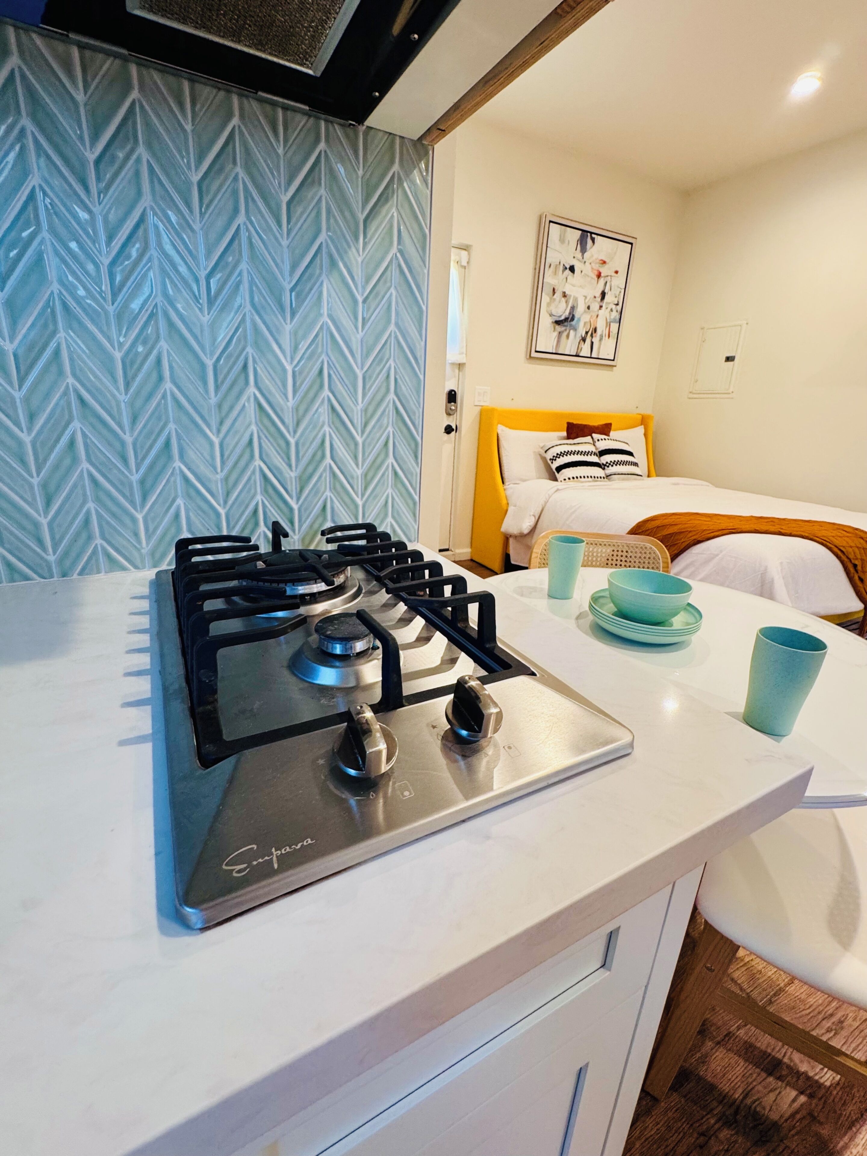 A kitchen with a gas stove, a white countertop, and a bed with a yellow headboard in the background.