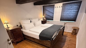 Un dormitorio con piso de madera, cama con ropa blanca, mesita de noche con lámpara, ventana con persianas y radiador.