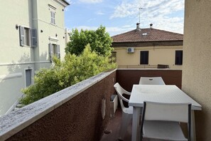 Un balcon avec une table et des chaises blanches, un mur en pierre et une vue sur les bâtiments voisins.