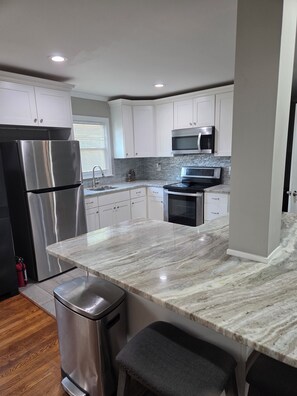 A modern kitchen with white cabinets, stainless steel appliances, a marble countertop, and a stainless steel trash can.