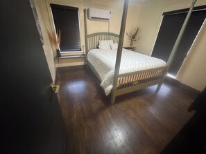 A bedroom with a wooden floor, a bed with a headboard, a window with blinds, and an air conditioning unit on the wall. - Pet Friendly  Sleeps 6  Fenced Yard  Patio (Elk City)