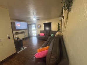 A living room with a dark brown leather couch, a flat-screen TV mounted on the wall, a ceiling fan, and a door leading outside. - Pet Friendly  Sleeps 6  Fenced Yard  Patio (Elk City)
