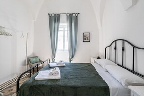 Ein Schlafzimmer mit einem Bett, einem Sessel, einem Nachttisch, einem Fenster mit Vorhängen und einem Wandbild.