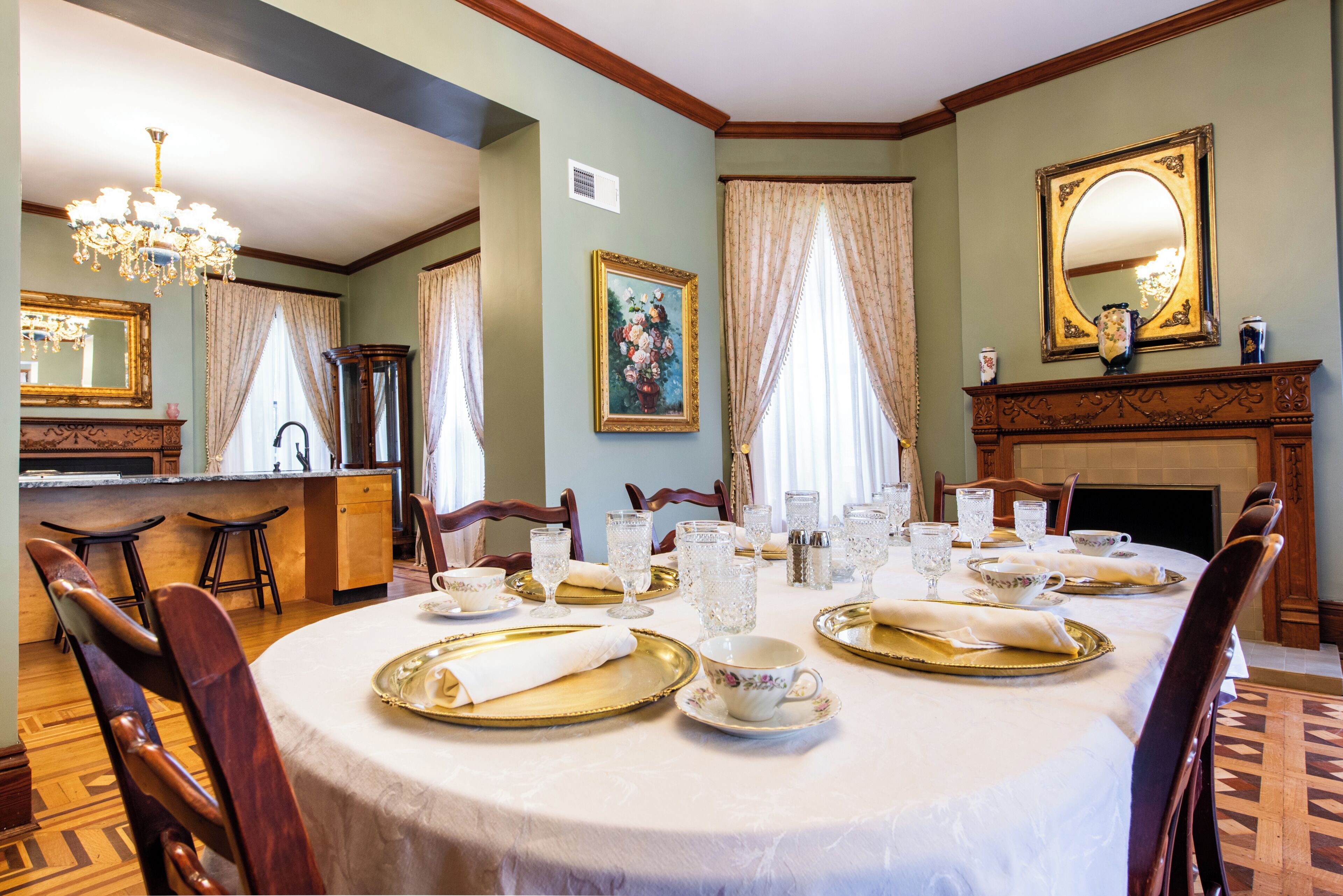 A dining room with a round table set for a meal, chairs, a chandelier, a fireplace, and a mirror on the wall.