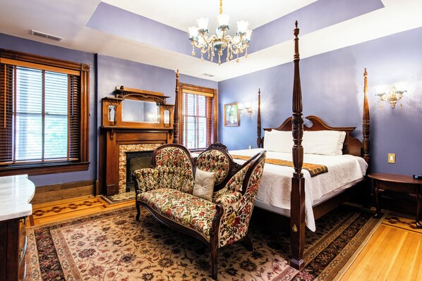 A bedroom with a four-poster bed, a floral armchair, a fireplace, and a large window with shutters.