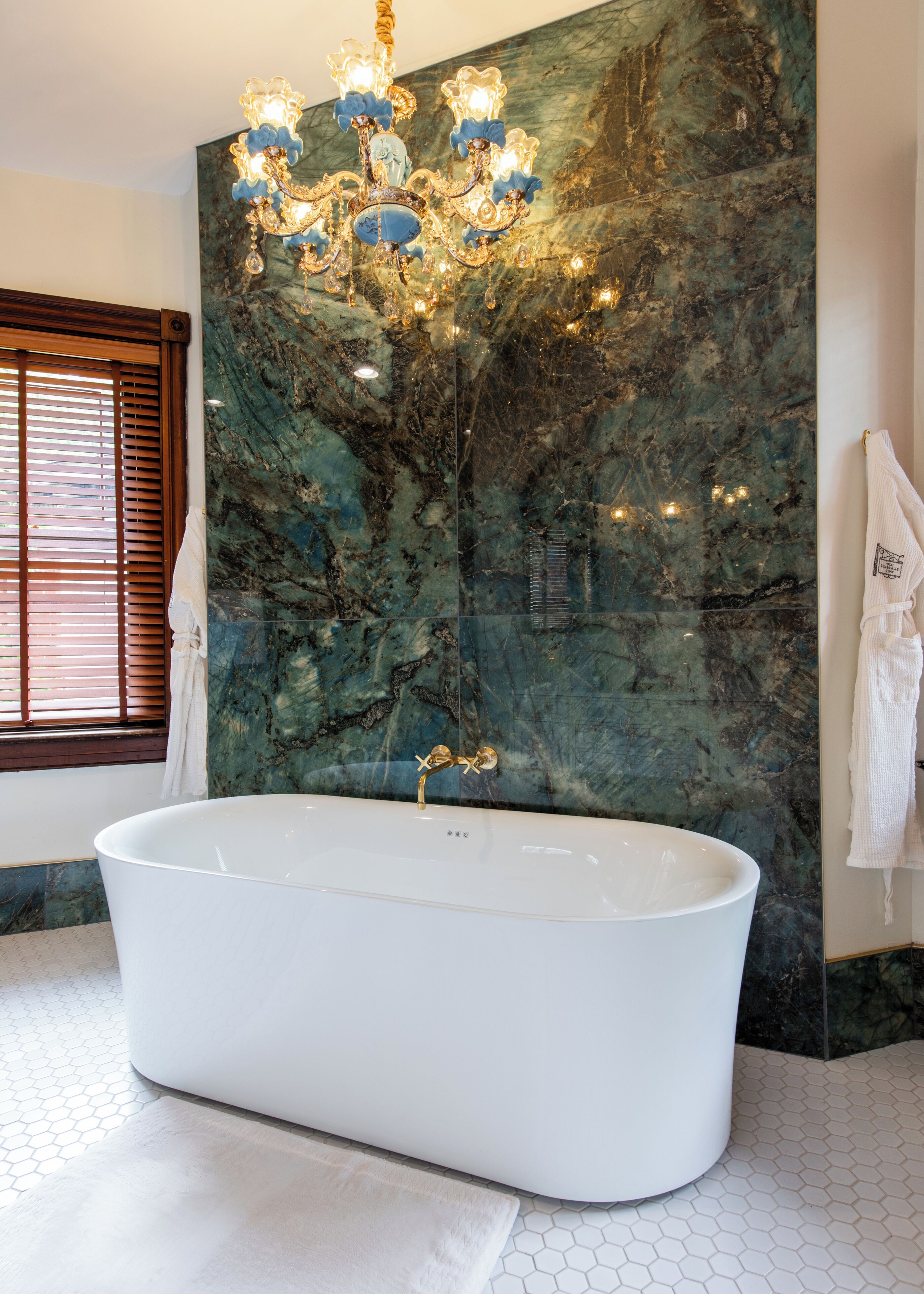 A bathroom with a freestanding bathtub, a marble wall, a chandelier, and a window with blinds.