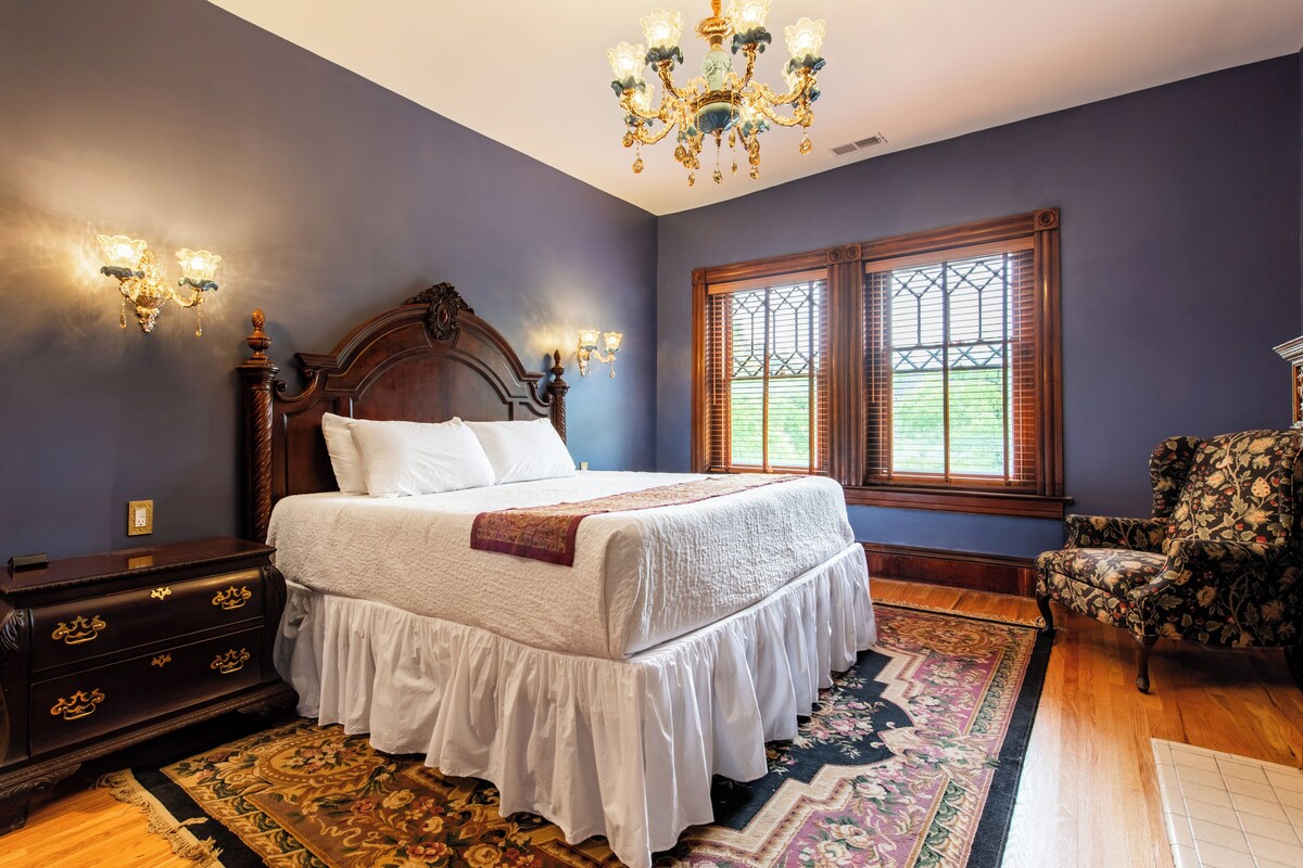 Une chambre à coucher avec un grand lit, une table de chevet en bois, une chaise, un lustre et une fenêtre avec des volets.