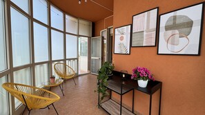 Ein Zimmer mit großen Fenstern, einem gelben Sessel, einem kleinen Tisch, einer Konsole mit einer Topfpflanze und gerahmten Bildern an der Wand.
