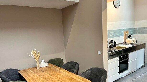 A dining area with a wooden table and four chairs, a kitchen with white cabinets and a black stove, and a clock on the wall. - Beautiful city apartment (Würzburg)