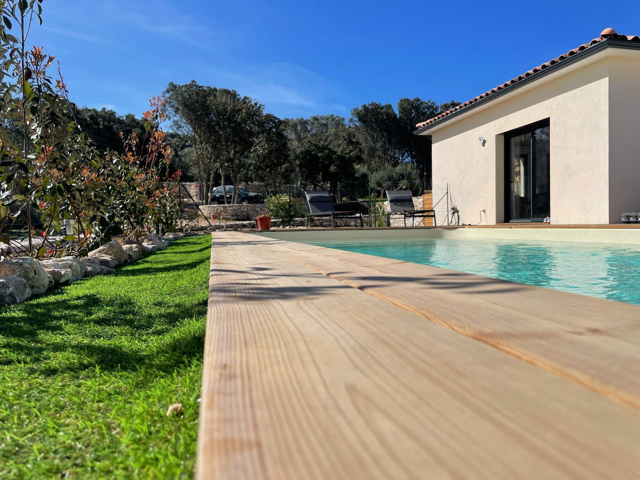 Una piscina con bordo in legno, un edificio moderno con ampie vetrate e un muro in pietra sullo sfondo.