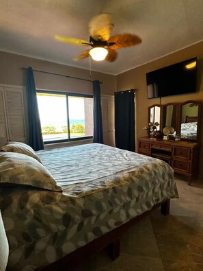Ein Schlafzimmer mit einem Bett, einem Deckenventilator, einem Schminktisch, einem Fernseher und einem Fenster mit Vorhängen.