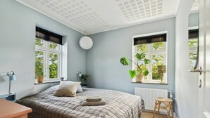 A bedroom with a bed, two windows, a radiator, a bedside table, a lamp, and a potted plant. - Big holiday home in retreat style (Millinge)
