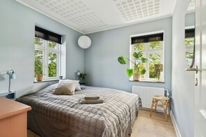 Ein Schlafzimmer mit einem Bett, zwei Fenstern, einer Heizung, einem Nachttisch, einer Lampe und einer Topfpflanze.