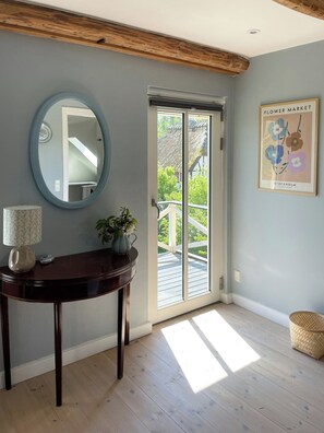 Ein Zimmer mit Holzboden, einem runden Spiegel, einem Tisch mit einer Vase, einem gerahmten Bild und einer Glasschiebetür, die auf eine Terrasse im Freien führt.