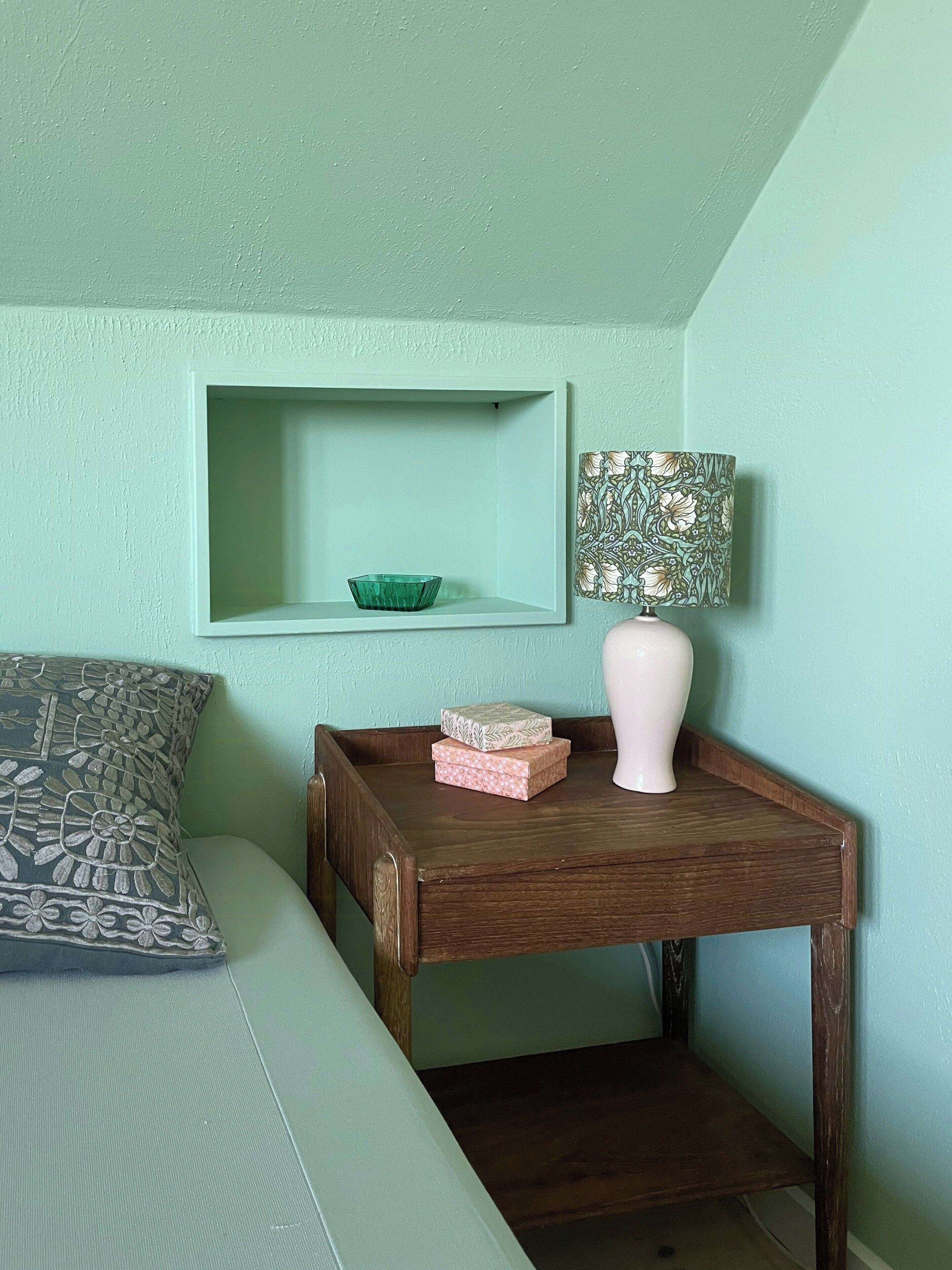 A room with a teal wall, a wooden nightstand with a lamp, a green bowl, and patterned boxes.